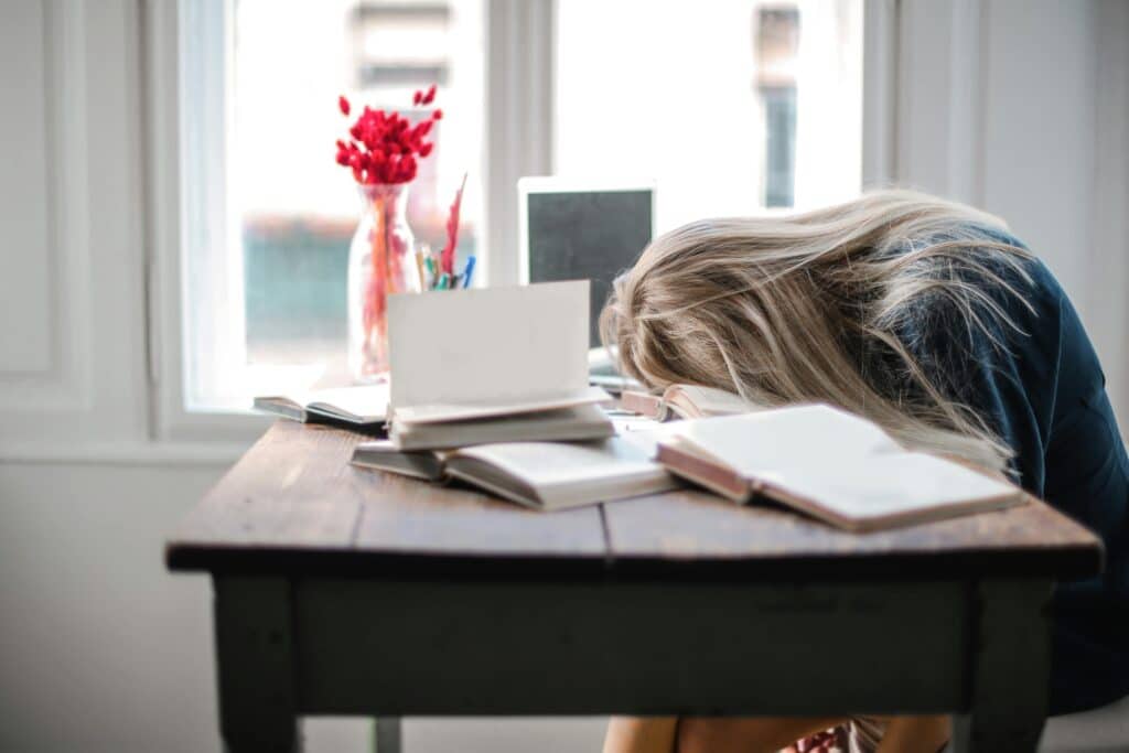 A stressed woman with long blonde hair rests her head on a wooden desk cluttered with open books and notebooks, appearing overwhelmed. A laptop and a vase with red flowers sit near a window in the background. A visual representation of the struggle to stop procrastinating and stay productive. Photo by Andrea Piacquadio: https://www.pexels.com/photo/woman-leaning-on-table-3767411/